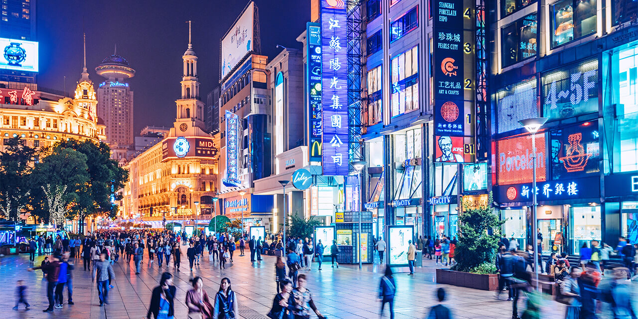 A busy city street in Shanghai, China at night, crowded with pedestrian traffic and illuminated by neon signs and building lights
